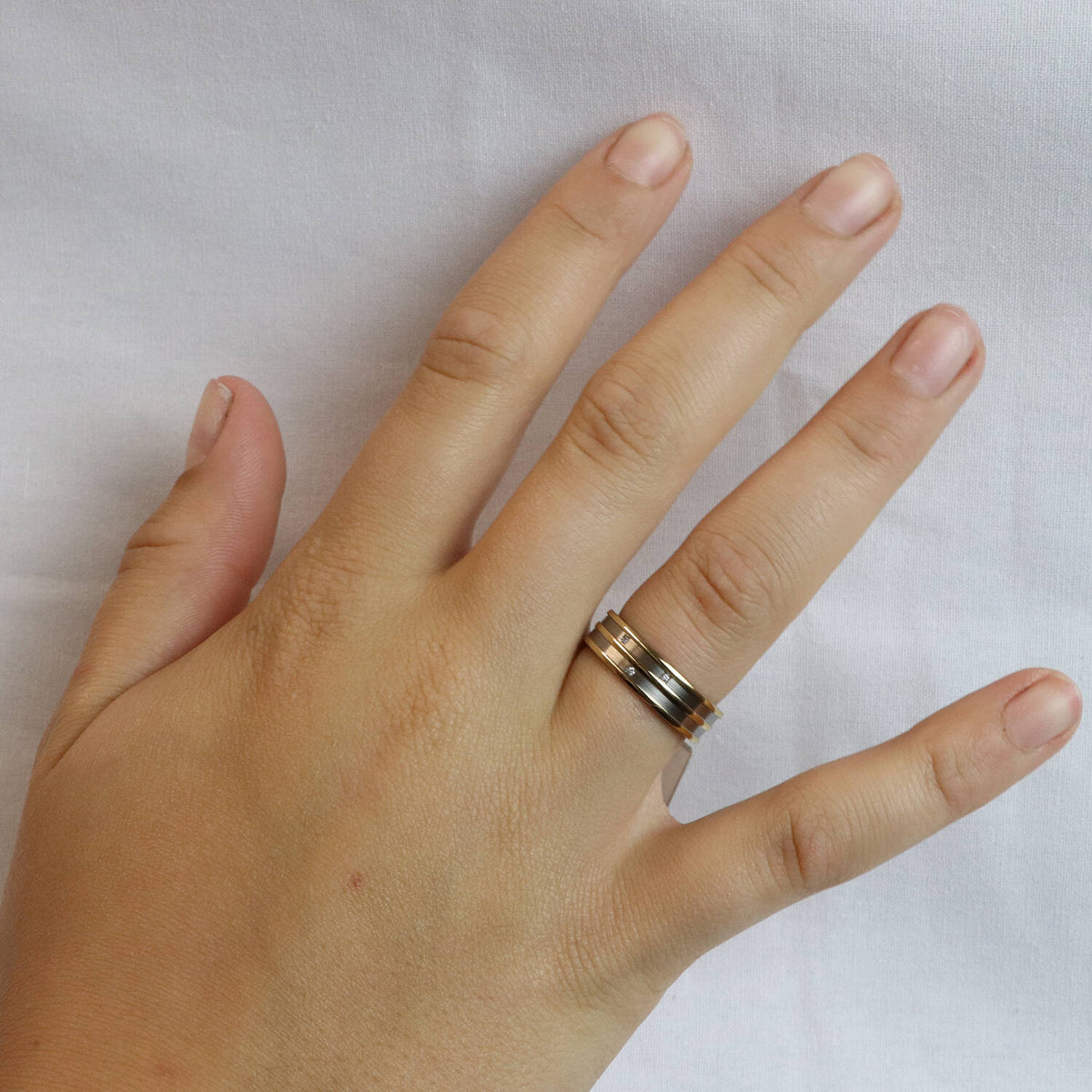 Een linkerhand met korte, natuurlijke nagels rust op een wit oppervlak. De ringvinger is voorzien van twee gestapelde metalen ringen: een chique Boccia Titanium Ring Bicolor dia 3/0.015 - 0151-02 en een eenvoudige zilveren band.