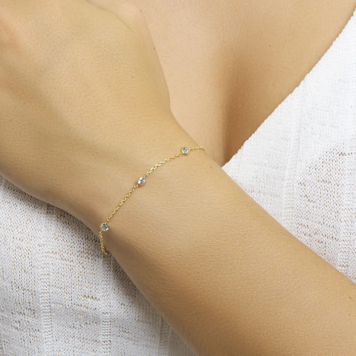 Een close-up van de pols van een vrouw met de Van der Kooij Collectie armband zirkonia 17 + 2 cm 3 micron zilver verguld (geel), versierd met kleine ronde zirkonia steentjes, afgezet tegen een wit gestructureerd bovenstuk.