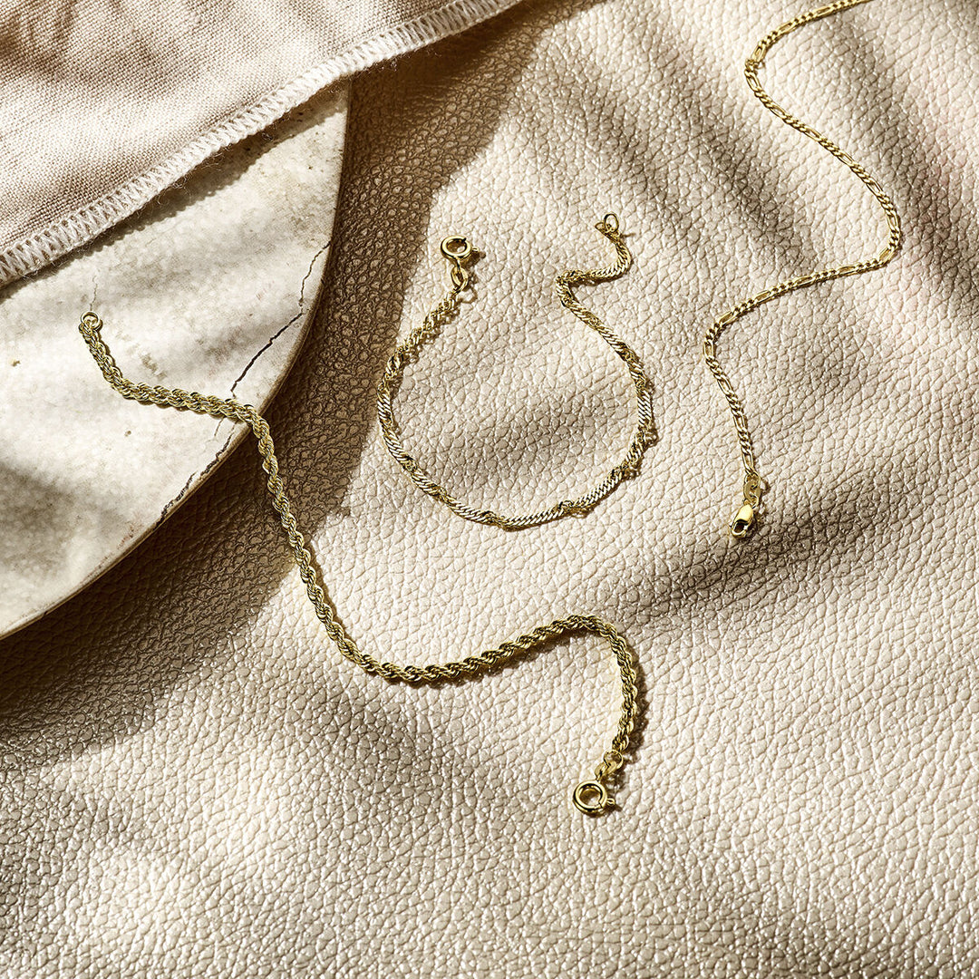 De armband figaro 1,8 mm 14K geelgoud van Van der Kooij Collectie wordt getoond op een crèmekleurig oppervlak met textuur, naast een beige doek en een keramische schaal, alles verlicht door zacht natuurlijk zonlicht.