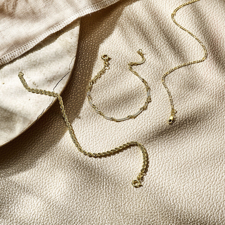 De armband figaro 1,8 mm 14K geelgoud van Van der Kooij Collectie wordt getoond op een crèmekleurig oppervlak met textuur, naast een beige doek en een keramische schaal, alles verlicht door zacht natuurlijk zonlicht.