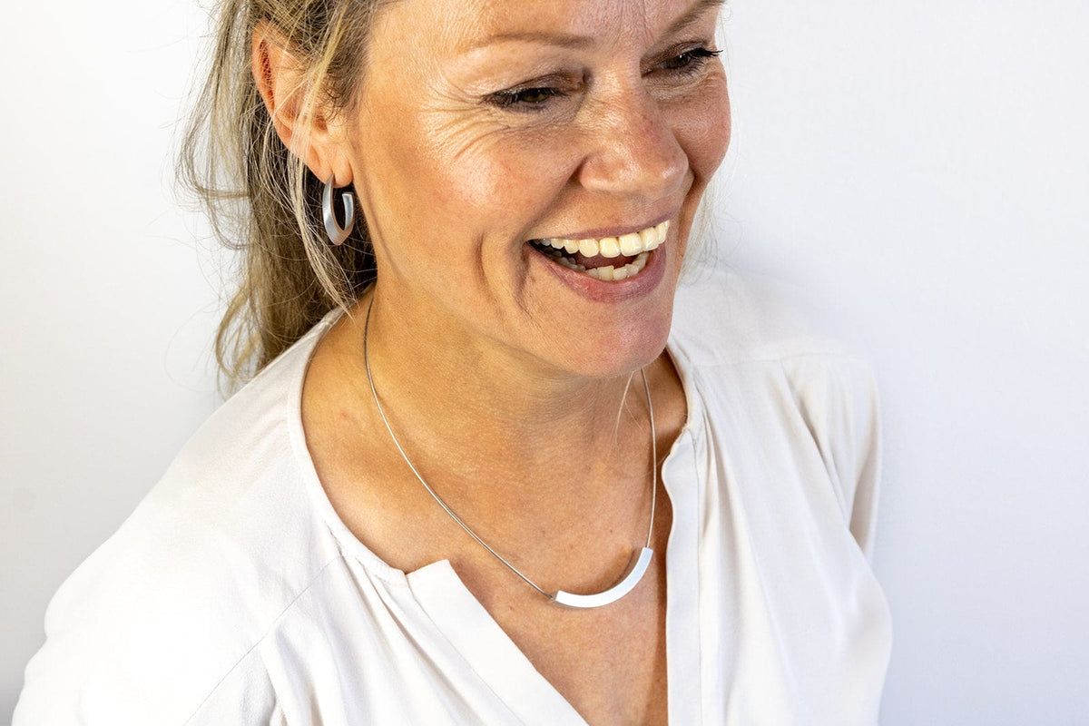 Een lachende vrouw in een lichte blouse draagt bijpassende geometrische oorbellen en een CLIC Creations Collier Bogen Goud|Geel C74G. Haar lichtbruine haar is naar achteren gebonden en ze staat tegen een effen witte achtergrond.