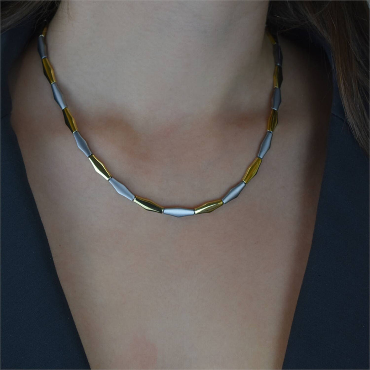 Een close-up van een persoon die de Boccia Titanium Collier Bicolor 08047-02 van Boccia draagt, met afwisselend gouden en zilveren langwerpige kralen, gestyled met een donkere top of blazer.