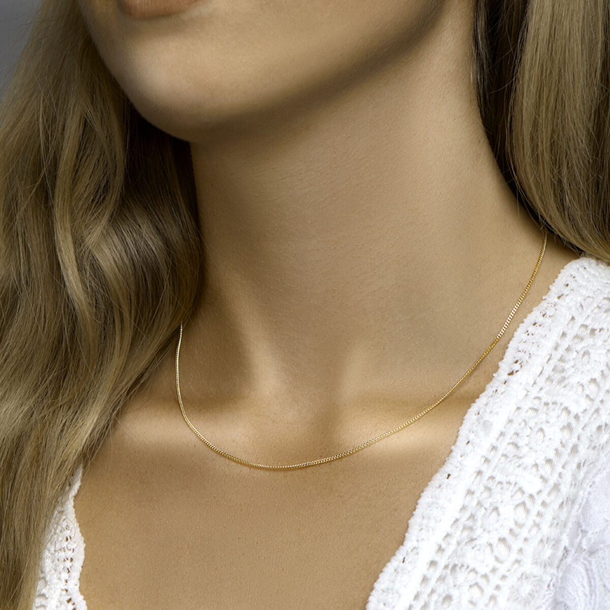 Een vrouw met lang blond haar draagt een Van der Kooij Collectie collier collier gourmette 4-zijdes geslepen 1,4 mm 3 micron zilver verguld (geel) en een wit kanten topje. De focus ligt op haar hals, waardoor het collier mooi uitkomt.