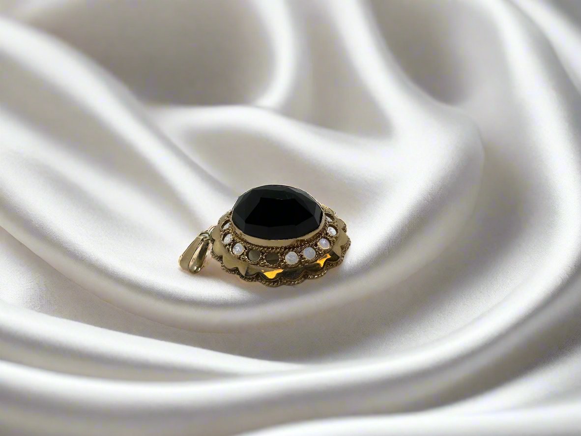 Op gladde witte zijde ligt een sierlijke 14 karaat gouden ring van Juwelier van der Kooij, met een grote zwarte edelsteen omringd door parelachtige accenten, wat doet denken aan een vintage collectie.