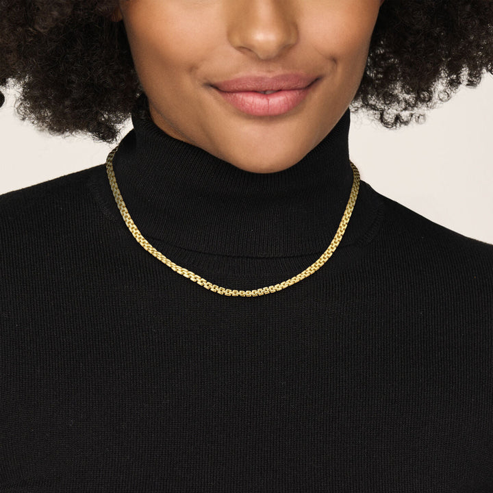 Een vrouw met krullend haar en natuurlijke make-up draagt een zwarte coltrui en het Van der Kooij Collectie collier panter 5 mm 43 cm in 14K geelgoud, terwijl ze zachtjes lacht tegen een effen achtergrond.