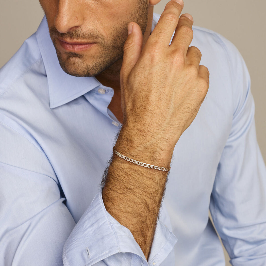 Een man in een lichtblauw overhemd laat zijn kin op zijn hand rusten, terwijl hij de Van der Kooij Collectie armband gourmette met balkje 3,8 mm 21 cm zilver wit showt. Zijn gezicht is deels zichtbaar tegen een neutrale achtergrond, waardoor deze stijlvolle heren accessoire extra aandacht krijgt.