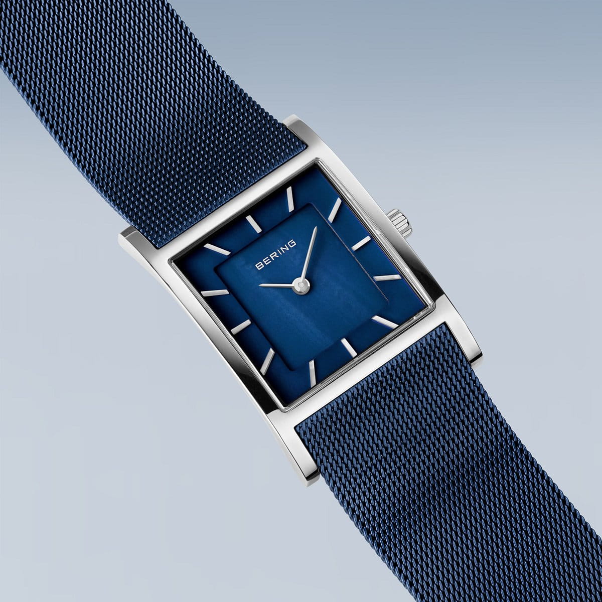 Het Bering dameshorloge 10426-307-S heeft een rechthoekig ontwerp met een blauwe wijzerplaat en meshband. Het bevat zilveren uurmarkeringen, wijzers en merknaam, afgezet tegen een lichtblauwgrijze achtergrond met kleurverloop. De zilveren kast wordt beschermd door duurzaam saffierglas en wordt aangestuurd door quartz uurwerk.