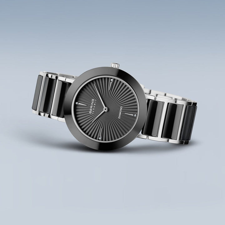 De Bering 11435-702 dameshorloge heeft een zwarte en zilverkleurige metalen band en een ronde zwarte wijzerplaat met radiale lijnen en minimalistische indexen, afgebeeld tegen een neutraal lichtverloop.