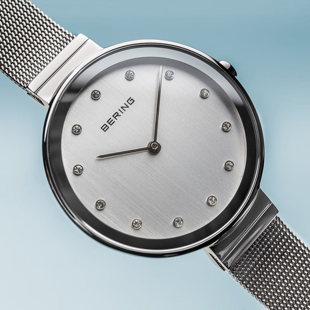 Het Bering 12034-000 Dameshorloge heeft een minimalistisch design op een blauwe achtergrond met een geborsteld metalen wijzerplaat, kristallen uurmarkeringen, zwarte wijzers en meshband. Verbeterd door quartz-uurwerk en beschermd door duurzaam saffierglas.