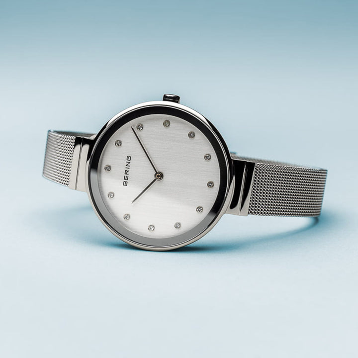 Het elegante Bering 12034-000 horloge heeft een minimalistisch design met een ronde wijzerplaat op een zilveren mesh band, subtiele uurmarkeringen, slanke wijzers tegen een lichte achtergrond onder duurzaam saffierglas, aangestuurd door quartz-uurwerk. Het rust op een lichtblauw oppervlak.