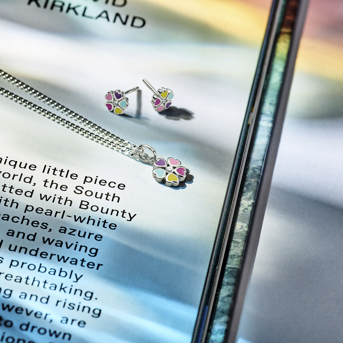 De Van der Kooij Collectie hanger bloem zirkonia zilver gerhodineerd ketting en bijpassende oorknopjes schitteren met zirkonia-accenten, weergegeven op een open boek badend in zacht regenboogkleurig licht.