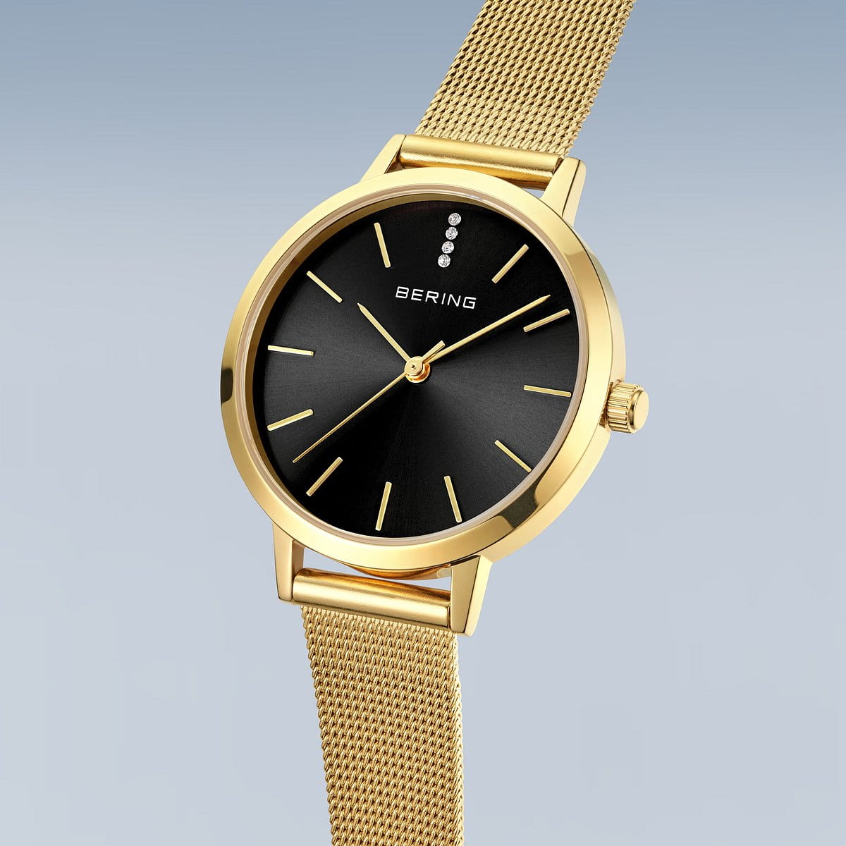 De Bering dames horloge zwarte wijzerplaat 13434-332 heeft een gouden kast, zwarte wijzerplaat met minimalistische markers, drie kristallen op 12 uur, een goudkleurige Milanees band en saffierglas op een lichtverloop achtergrond.