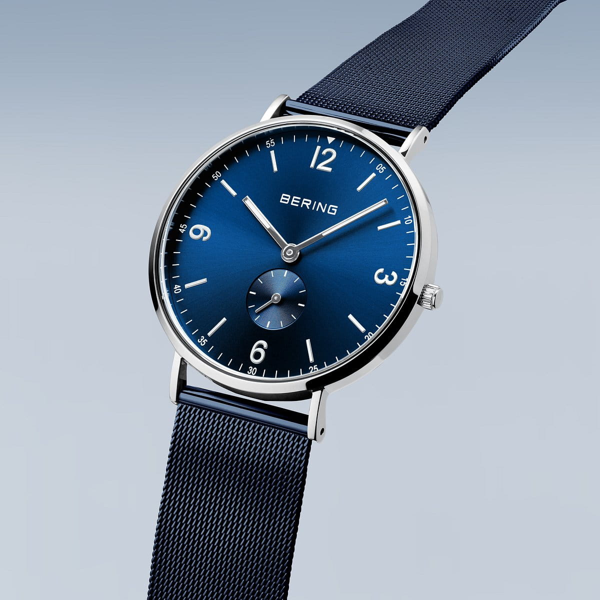 Het Bering heren horloge blauw 14040-307 heeft een blauwe wijzerplaat, zilverkleurige uurmarkeringen en een blauwe mesh band, met een kleine secondewijzer op 6 uur. Dit minimalistische Bering horloge is afgebeeld tegen een lichtblauwe achtergrond.