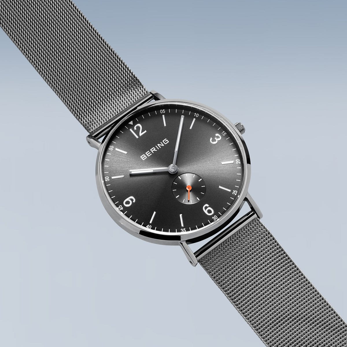 De Bering heren horloge grijs 14040-377 heeft een grijze mesh band, zwarte wijzerplaat, witte uurmarkeringen, zilverkleurige wijzers, een sub-dial boven 6 uur en een quartz uurwerk - allemaal van Bering - tegen een lichtblauwe achtergrond.