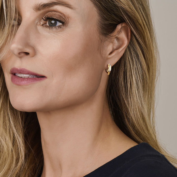 Een vrouw met blond haar en lichte make-up draagt een donker topje en Van der Kooij Collectie oorringen 1 micron zilver verguld (geel/rhodium), iets opzij kijkend tegen een neutrale achtergrond.