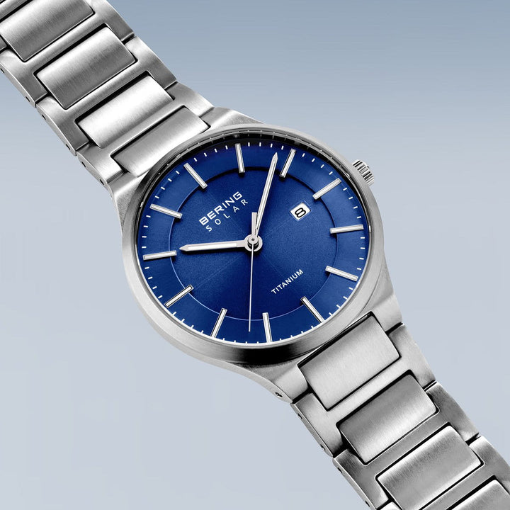 De Bering 15239-777 is een zilveren Herenhorloge met een gekoppelde titanium band en blauwe wijzerplaat met witte uurmarkeringen, een klein datumvenster, uur- en minutenwijzers. De wijzerplaat toont Bering en TITANIUM op een lichtblauwe achtergrond.