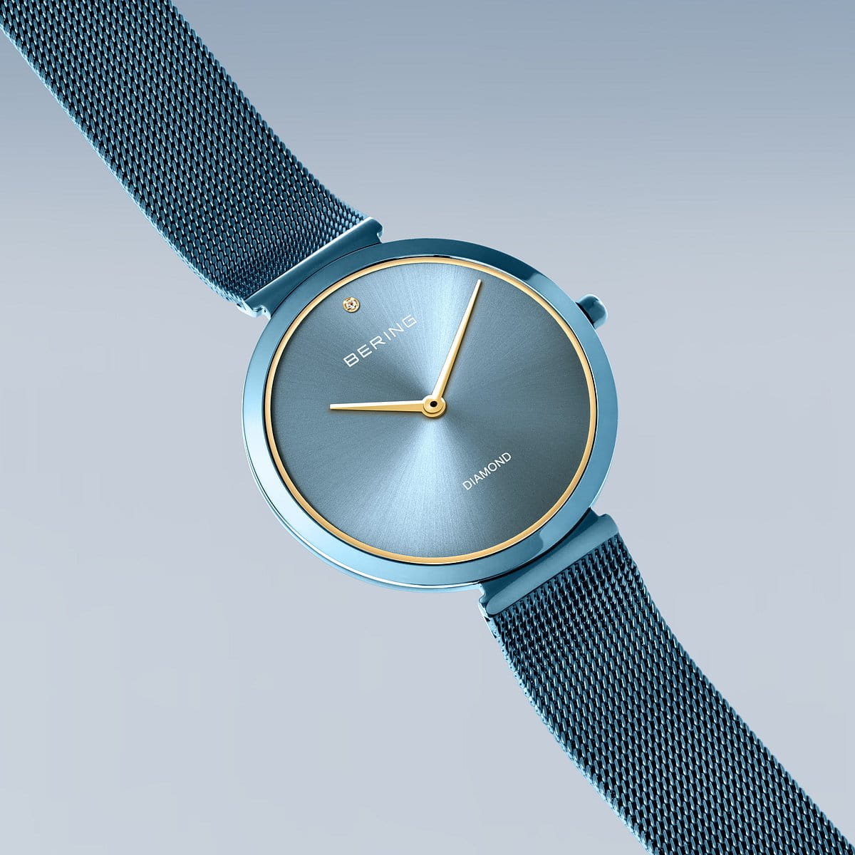 De Bering dames horloge blauwe wijzerplaat - 18132-Charity1 heeft een minimalistische blauwe wijzerplaat, goudomrande kast, eenvoudige gouden wijzers, een diamant op 12 uur en een blauwe RVS mesh horlogeband, weergegeven op een lichtblauwe achtergrond.