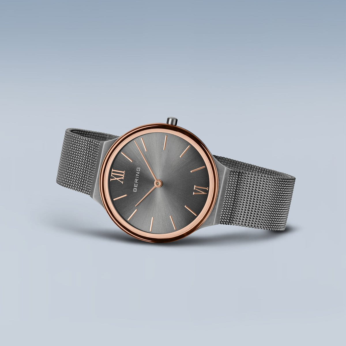 Het Bering dameshorloge 18434-369 heeft een roségouden kast, grijze wijzerplaat met Romeinse cijfers en grijze mesh band. Het is voorzien van duurzaam saffierglas en wordt elegant weergegeven op een lichtblauwe achtergrond.