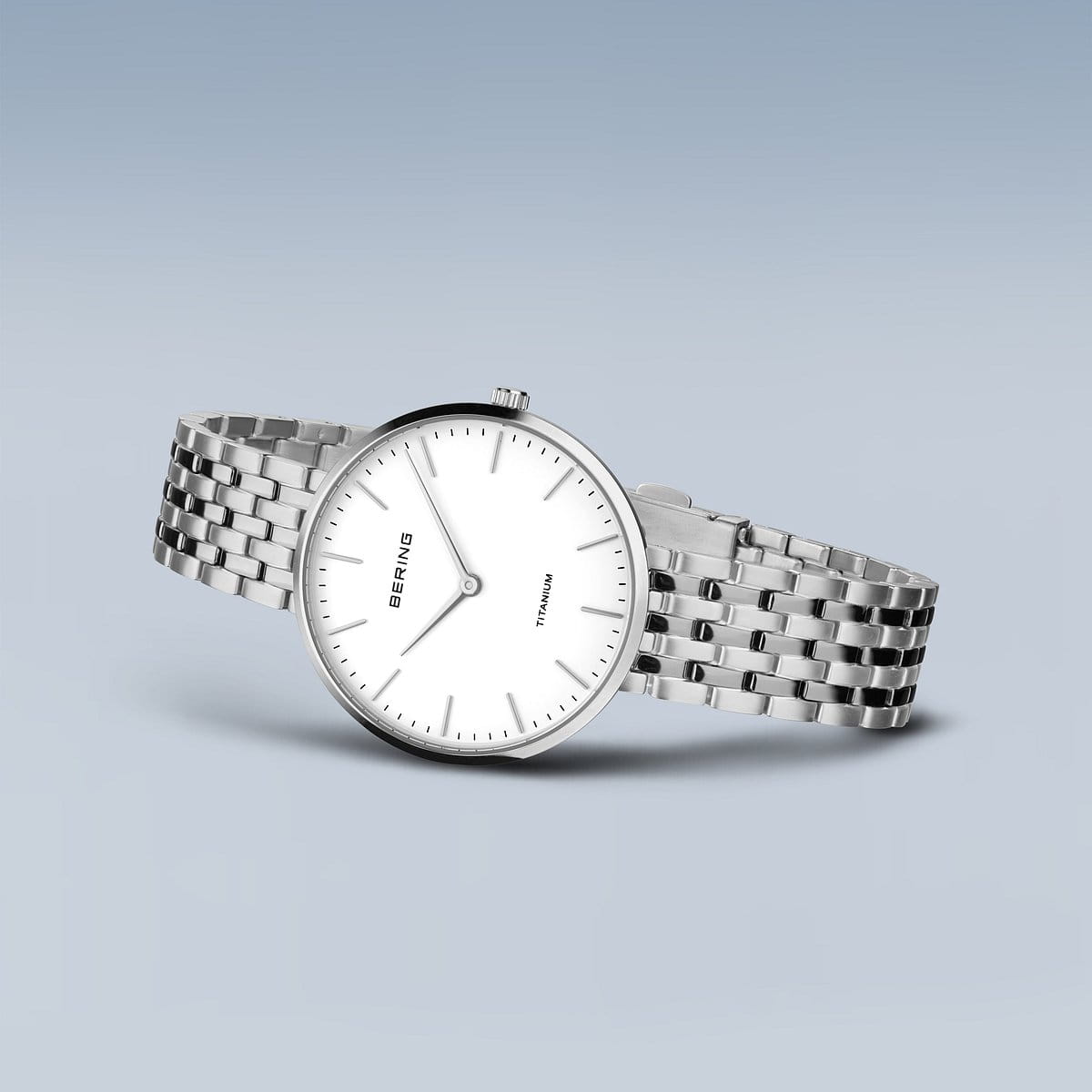 Het Bering dameshorloge 19334-004, met zijn witte wijzerplaat en slanke uurmarkeringen, rust op een lichtblauwe achtergrond. Dit strakke horloge heeft een gekoppelde metalen band en wordt beschermd door duurzaam saffierglas.