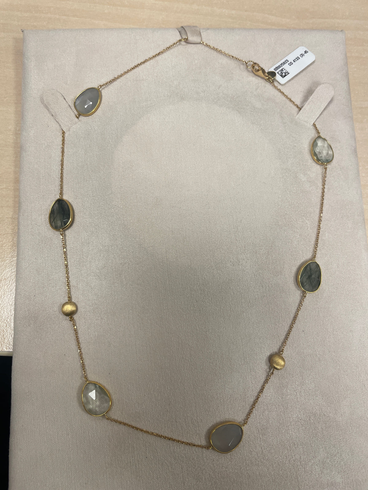 De Italo Bottene CO 4133 (3)-45 ketting is een delicaat, goudkleurig sieraad met ovale stenen in gedempte grijs- en groentinten, afgewisseld met kleine gouden kraaltjes, elegant tentoongesteld op een beige gewatteerd oppervlak.