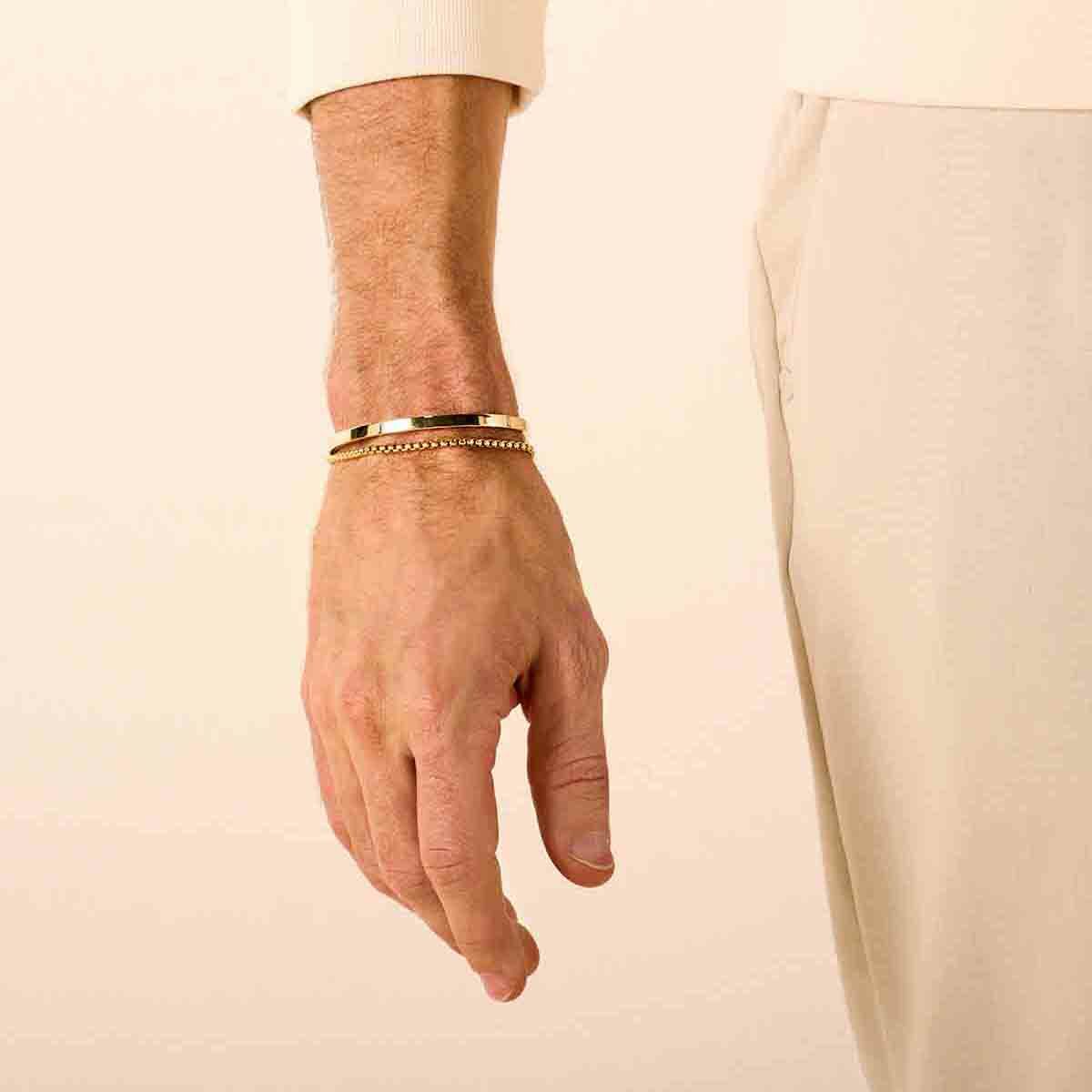 Een close-up toont een linkerhand en -pols met twee Van der Kooij Collectie Gouden armband heren venetiaans bol 2,5 mm 14K, met de drager in een crèmekleurige trui en beige broek tegen een neutrale achtergrond.