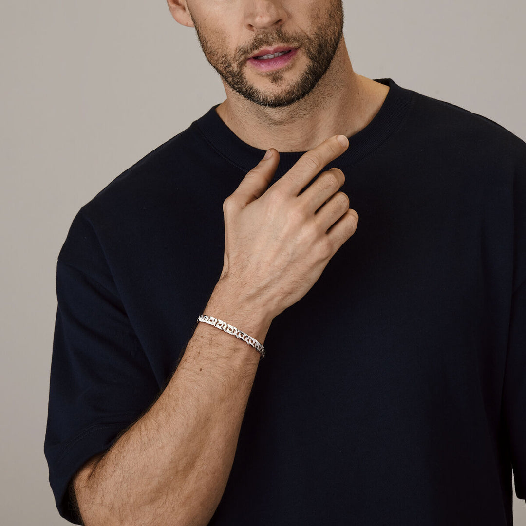 Een man met een bijgeknipte baard en een donkerblauw t-shirt poseert met zijn hand bij zijn gezicht en toont de armband van 6,5 mm zilverwit van Van der Kooij Collectie om zijn pols.