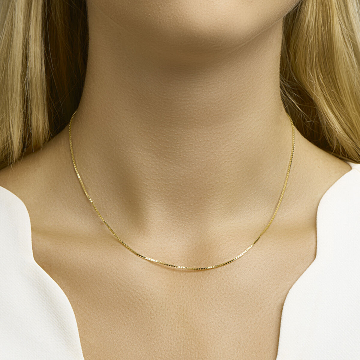 Een vrouw met lang blond haar draagt een Van der Kooij Collectie collier venetiaans 1,1 mm 14K geelgoud in een delicate Venetiaanse schakel, gecombineerd met een wit topje met V-hals, dat vanaf de schouders zichtbaar is.