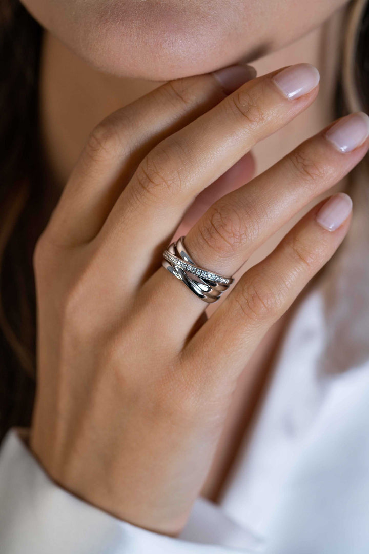 Een close-up van de hand met natuurlijke nagels van een vrouw, met gestapelde zilveren ringen, waaronder de Zinzi Ring zilver ZIR1790, vlak bij haar gezicht. Ze is gekleed in een witte outfit met lange mouwen.