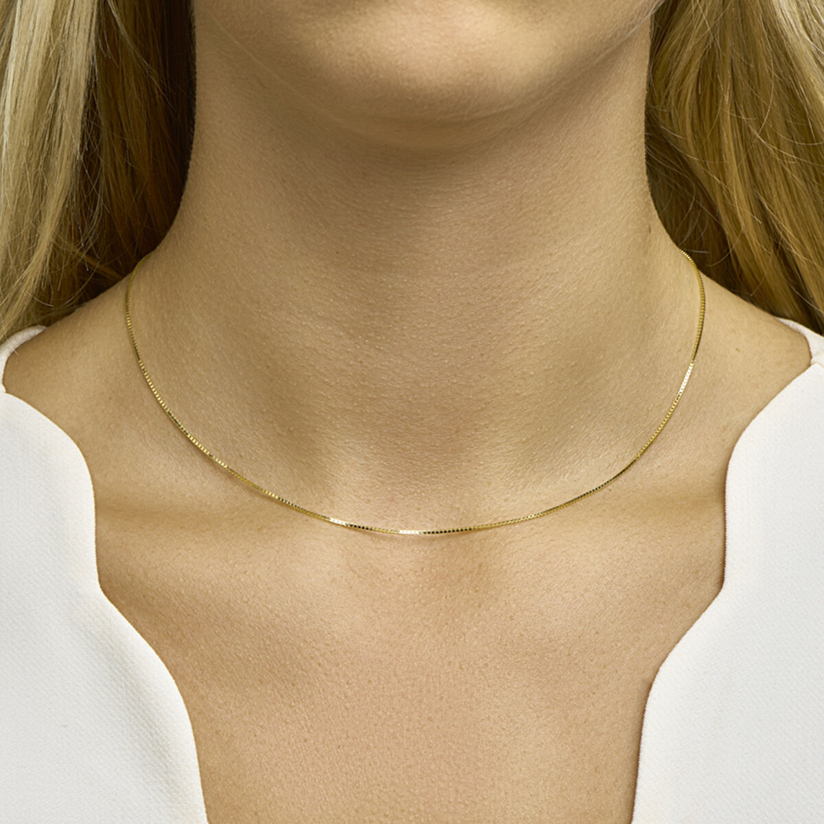 Een vrouw draagt het delicate Collier Venetiaans 0,8 mm 14K geelgoud uit de Van der Kooij Collectie, een dun gouden collier met een Venetiaanse schakel, dat haar blote nek siert boven een wit topje met V-hals.