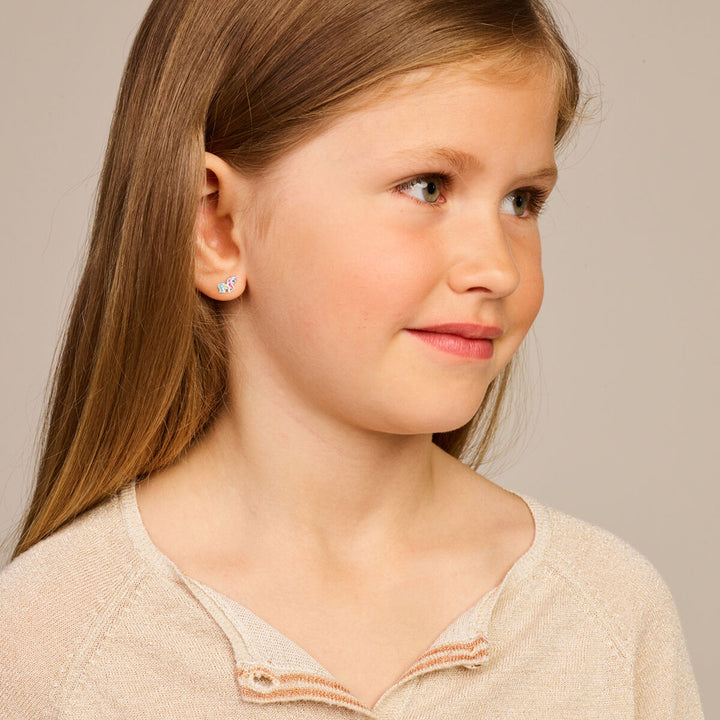 Een jong meisje met lang lichtbruin haar, draagt een beige trui en Van der Kooij Collectie oorknoppen eenhoorn zilver gerhodineerd, glimlacht zachtjes terwijl ze opzij kijkt tegen een neutrale achtergrond.