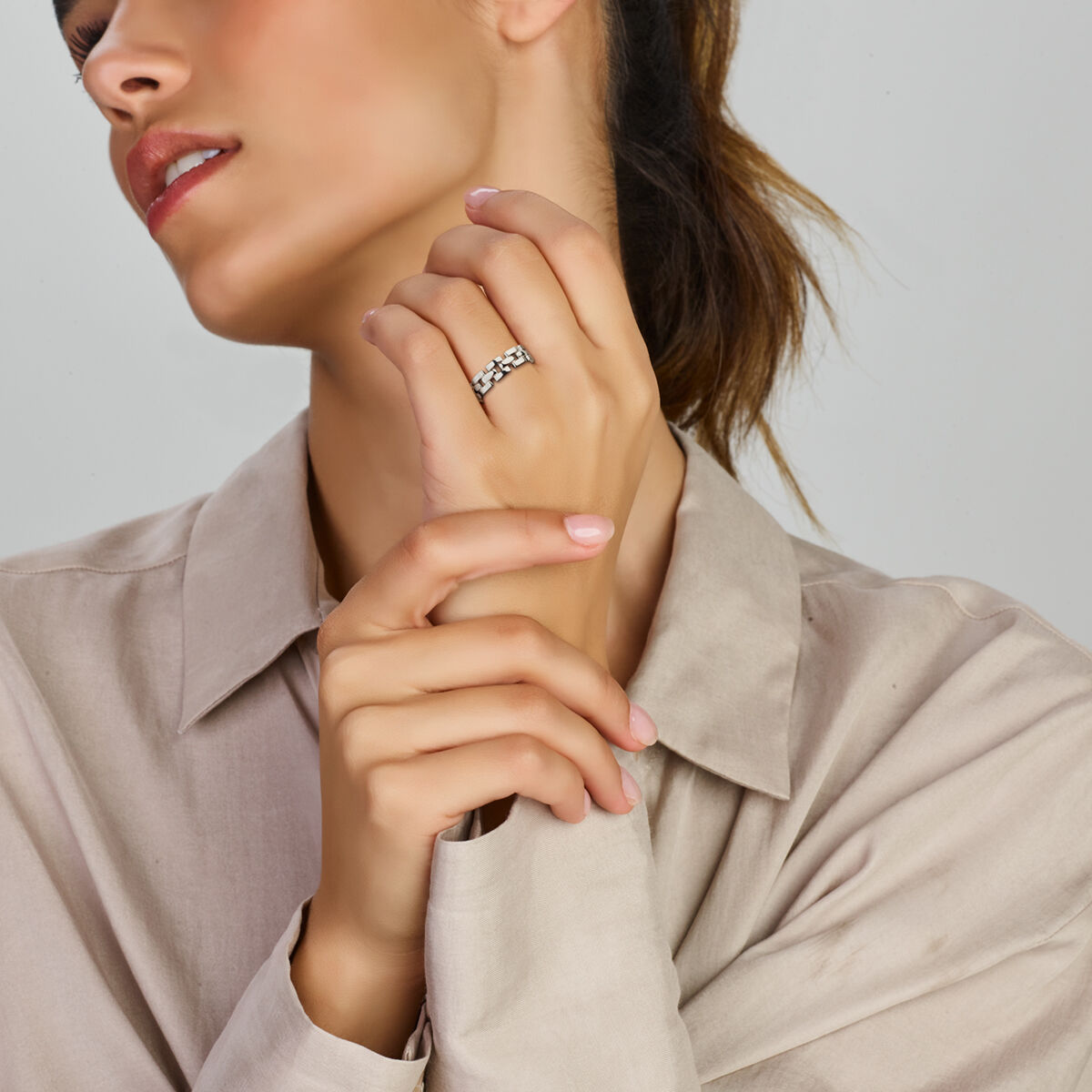 Een vrouw in een beige overhemd presenteert de Van der Kooij Collectie ring schakel zilver gerhodineerd met beide handen bij haar gezicht, haar hoofd iets gedraaid om haar lippen te accentueren terwijl de tijdloze schoonheid van de ring afsteekt tegen een neutrale achtergrond.