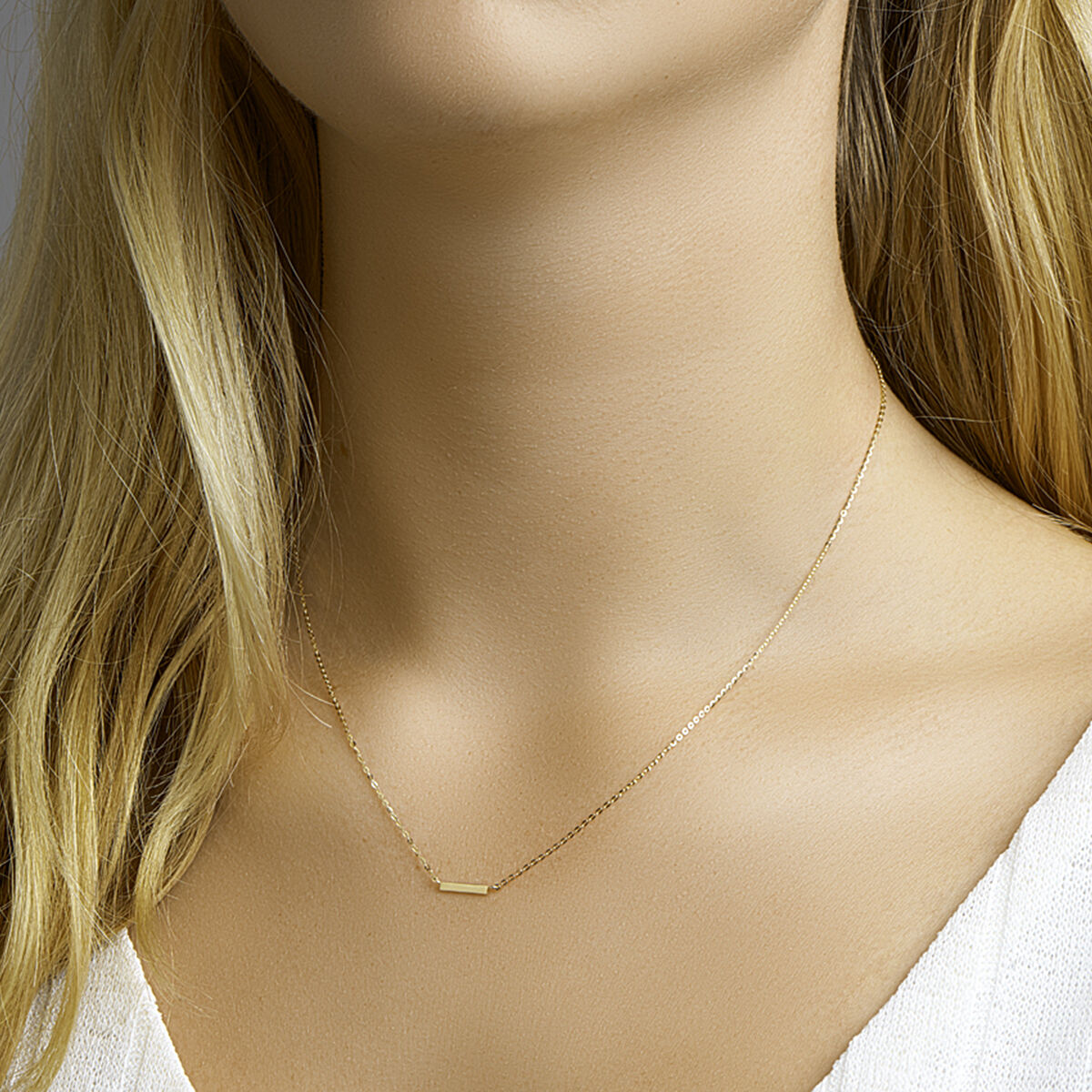 Een close-up van een vrouw met lang blond haar en een witte top, die het Van der Kooij Collectie collier balkje 41 + 4 cm van 14K geelgoud draagt, met een subtiel rechthoekig hangertje in ankerschakelontwerp.