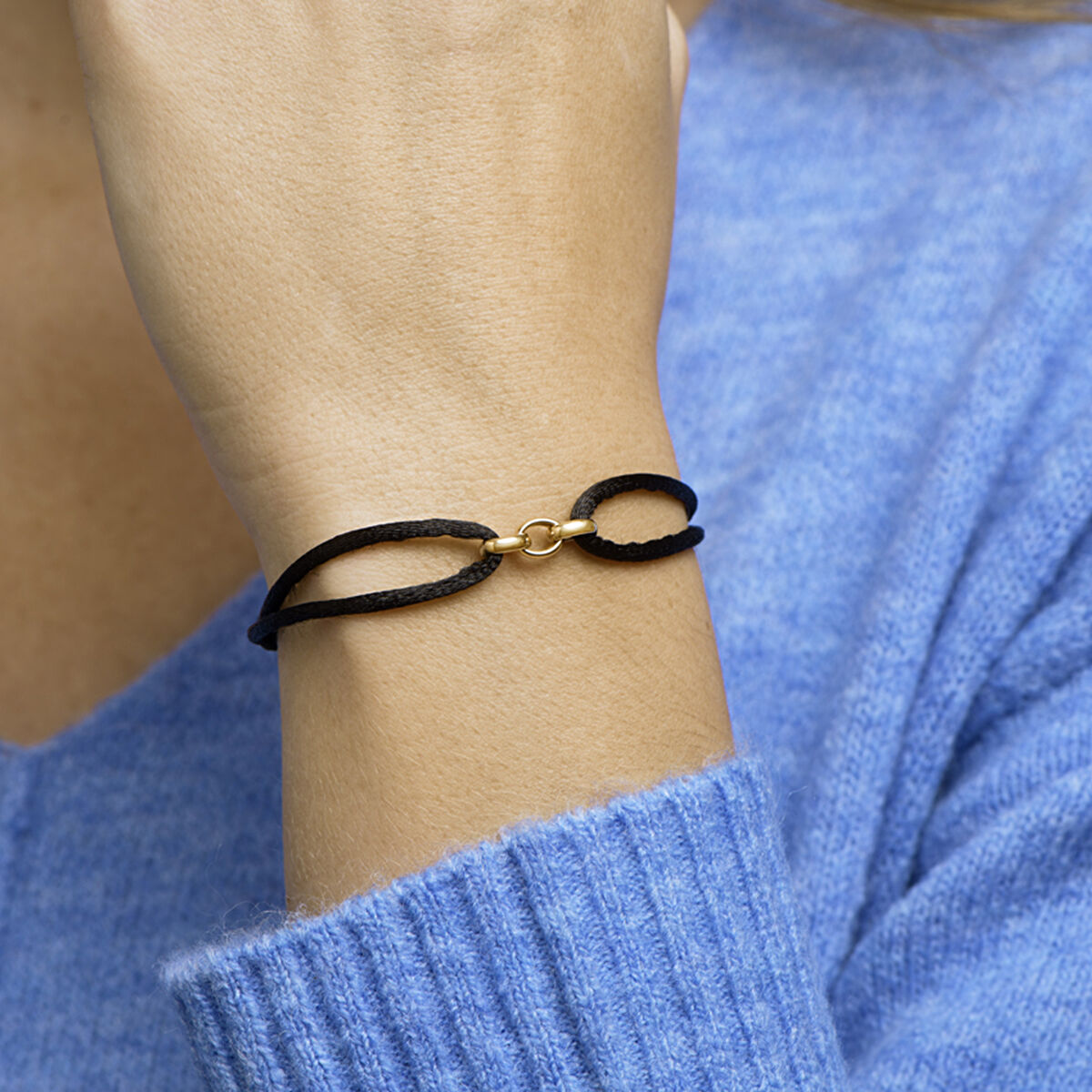 Een persoon in een blauwe trui showt een Van der Kooij Collectie Armband satijn 14K goud met rubber/nylon/leer - een zwarte koordarmband met een gouden detail - om zijn pols, de hand voorzichtig naar zijn gezicht geheven.