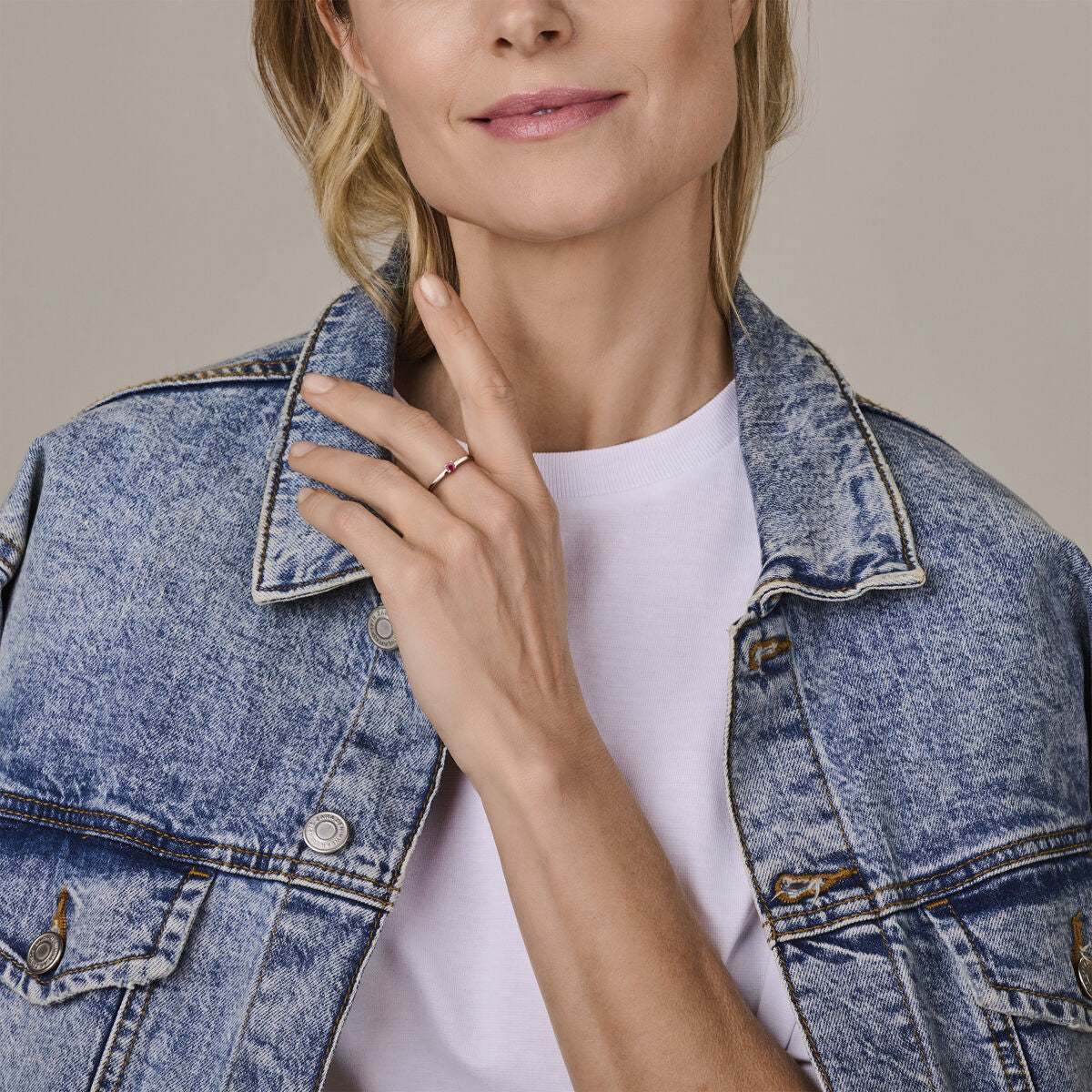 Een vrouw in een licht spijkerjasje over een wit T-shirt glimlacht zachtjes en toont de Van der Kooij Collectie ring met geboortesteen robijn juli zilver gerhodineerd aan haar linkerhand. De foto is bijgesneden bij haar neus om het sieraad te benadrukken.