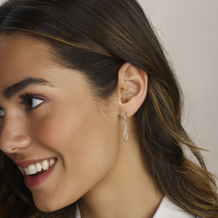 Een vrouw met golvend bruin haar lacht terwijl ze Van der Kooij Collectie oorhangers zirkonia brisur haak zilver gerhodineerd draagt. De foto belicht haar linkeroor, oorbel en een deel van haar witte topje.