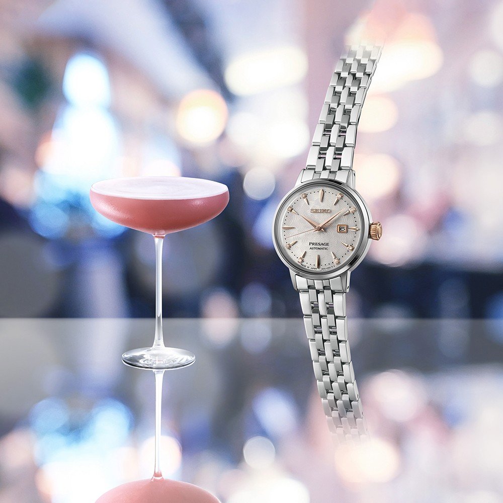 Een Seiko Presage dameshorloge SRE009J1 met een zilveren metalen schakelband staat naast een roze cocktail op een reflecterend oppervlak. Het Hardlex mineraalglas schittert en creëert een bokeh-effect met warme lichten en wazige vormen.