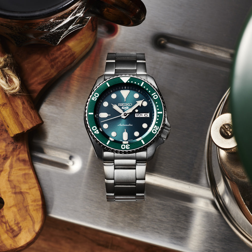 Een Seiko 5 Sports herenhorloge SRPD61K1 met een metalen band en groene lunette staat op een metalen oppervlak. Het automatisch horloge toont de dag en datum, vergezeld door een houten object en groene ketel, die een casual tafelbladscène vormen.