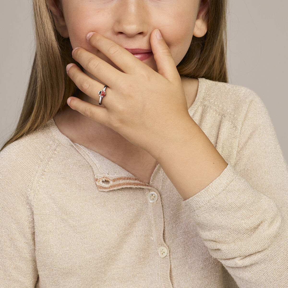 Een jong meisje met lang lichtbruin haar in een beige vest bedekt haar mond en glimlacht lichtjes. Aan haar ringvinger draagt ze de Van der Kooij Collectie ring lieveheersbeestje zilver gerhodineerd.