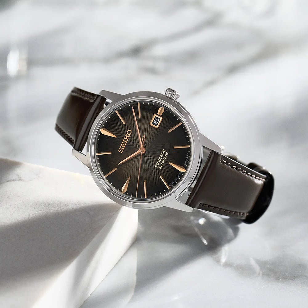 De Seiko Presage herenhorloge SRPJ17J1 heeft een bruine wijzerplaat, datumweergave, bruine leren band en zilveren kast. Dit automatische uurwerk staat op wit marmer, de elegantie wordt benadrukt door schaduwen op het Hardlex mineraalglas.