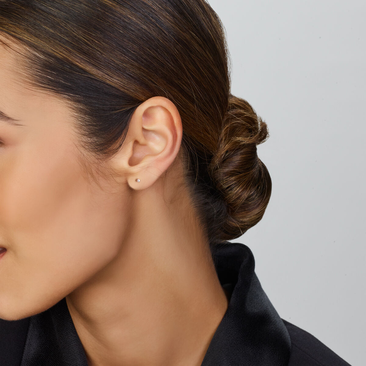 In profiel is een vrouw met donker haar in een nette knot te zien, gekleed in oorknoppen bolletje zilver gerhodineerd van Van der Kooij Collectie en een kledingstuk met zwarte kraag tegen een effen lichte achtergrond.