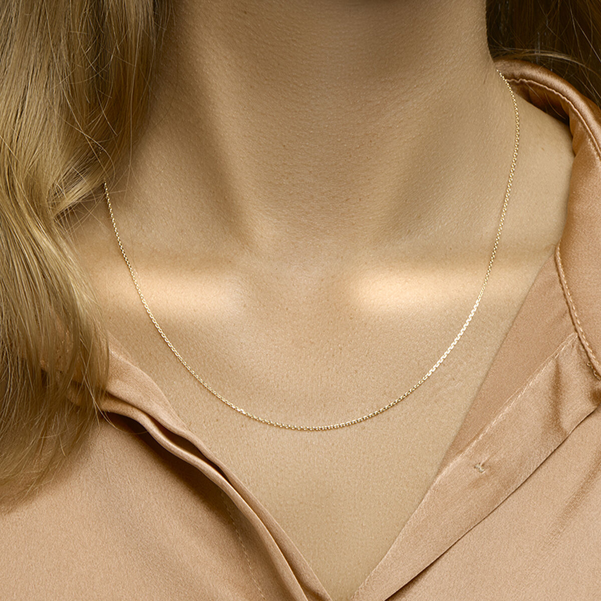 Een close-up toont een persoon die de Van der Kooij Collectie collier anker gediamanteerd 1,1 mm 41 + 4 cm 3 micron zilver verguld (geel) ketting draagt over een lichtbruine satijnen blouse; alleen de hals, het sleutelbeen en een deel van de blouse zijn zichtbaar.