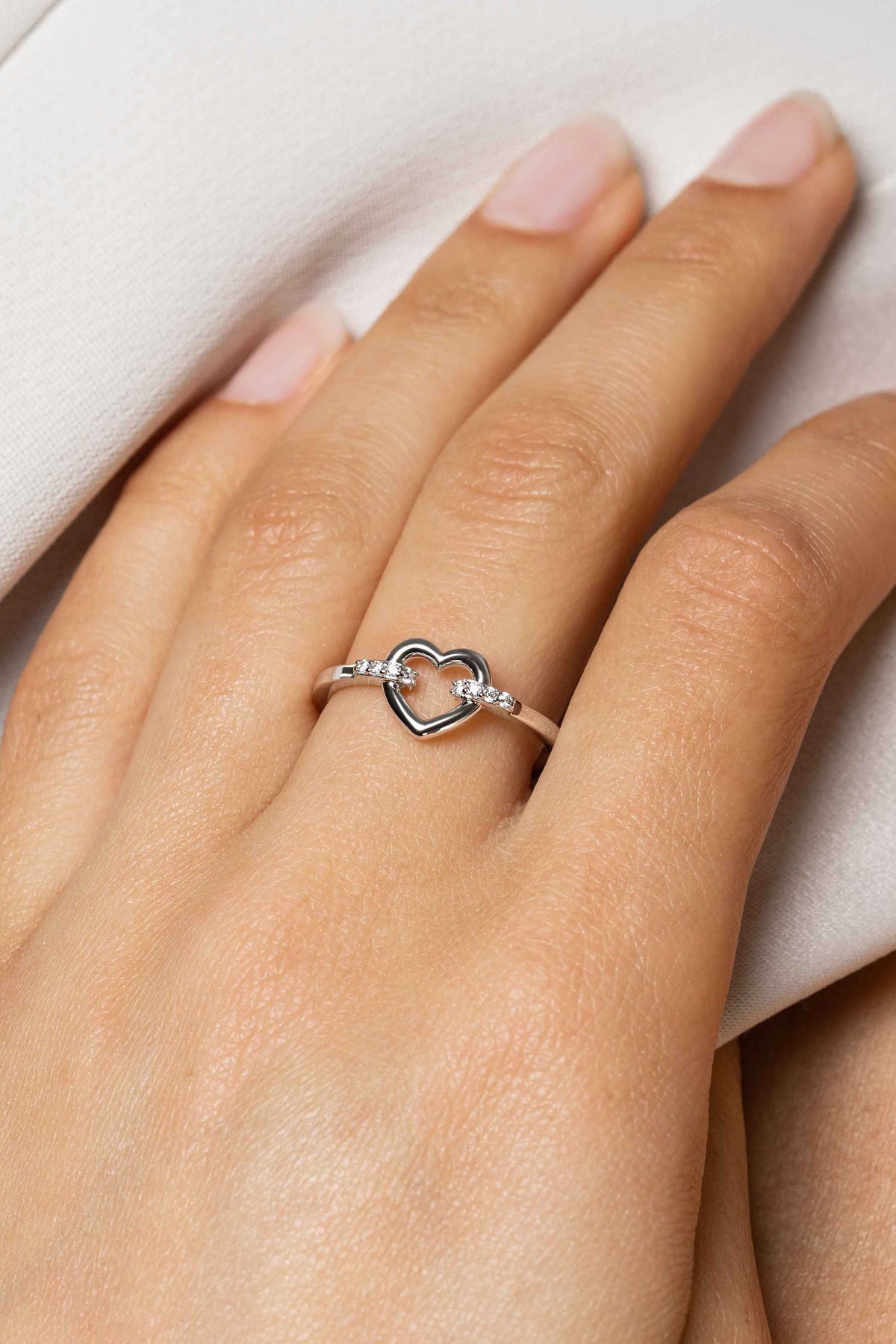 Een close-up van een hand die de Zinzi Ring Zilver Zir1755 van Zinzi draagt, met een open hart ontwerp met kleine witte zirkonia's aan elke kant, elegant gedragen aan de ringvinger tegen een witte stof.