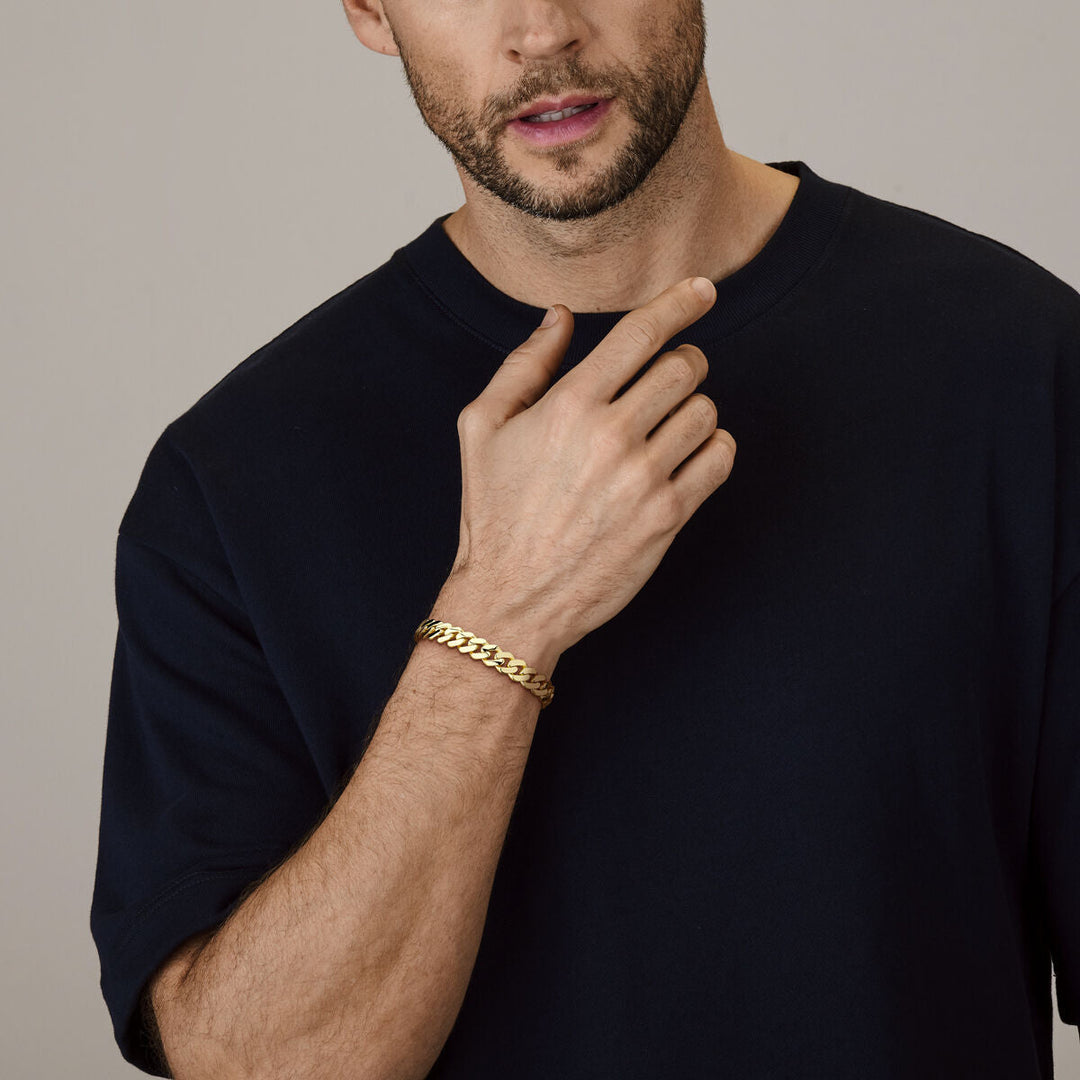 Een man in een donker t-shirt raakt zijn hals aan en toont de Van der Kooij Collectie armband massief gourmette 10 mm 21 cm in 18K geelgoud om zijn pols tegen een neutrale achtergrond.