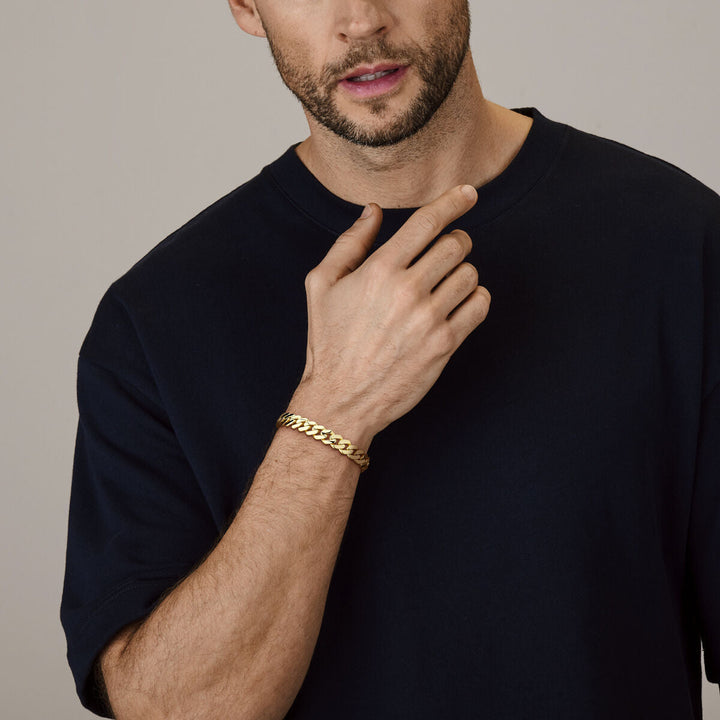 Een man in een donker t-shirt raakt zijn hals aan en toont de Van der Kooij Collectie armband massief gourmette 10 mm 21 cm in 18K geelgoud om zijn pols tegen een neutrale achtergrond.