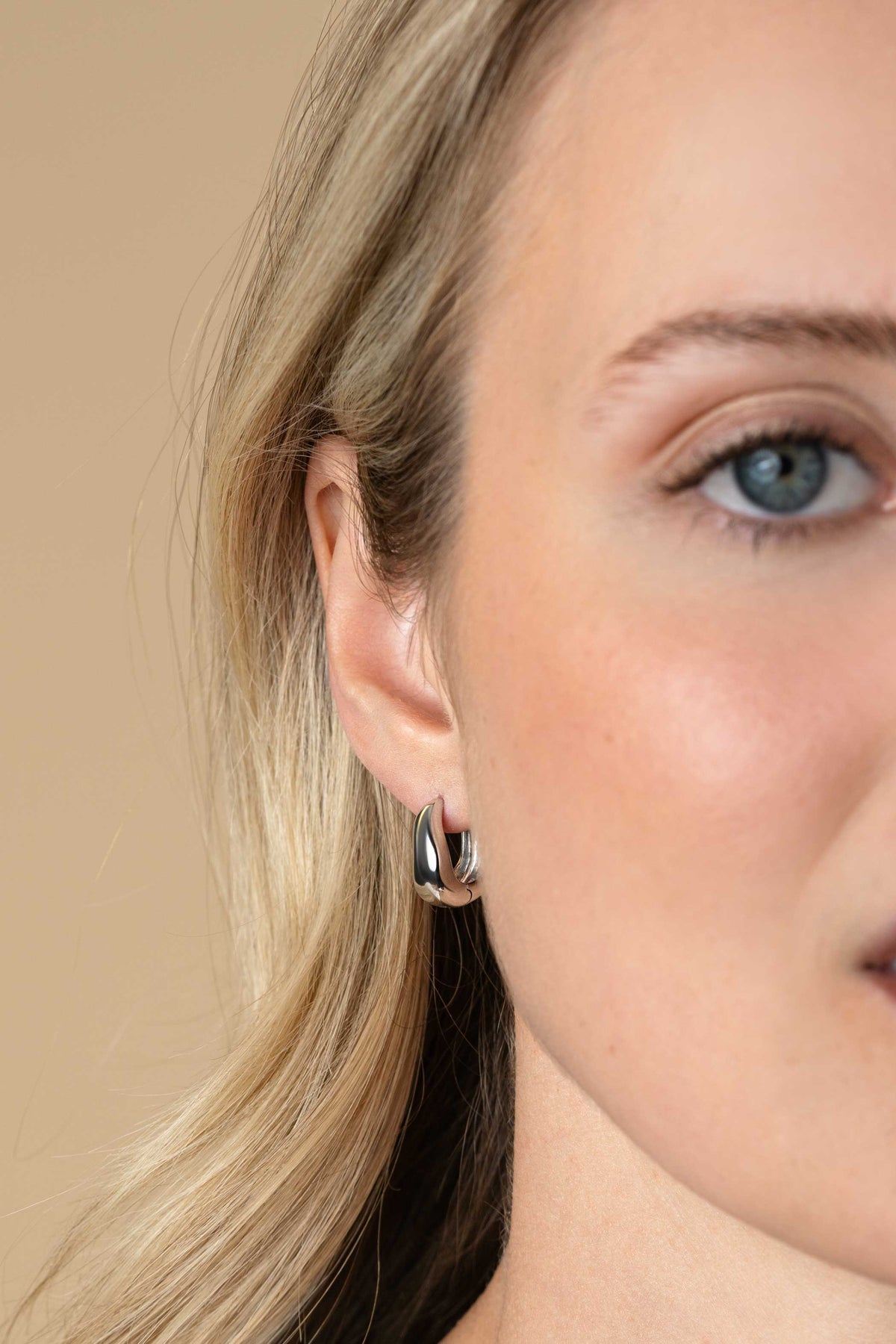 Close-up van een vrouw met blond haar en een lichte huid die Zinzi oorbellen zilver ZIO2609 van Zinzi draagt, tegen een neutrale beige achtergrond. Slechts een deel van haar gezicht en hoofd is te zien, waardoor de kleine, dikke zilveren oorbellen goed uitkomen.
