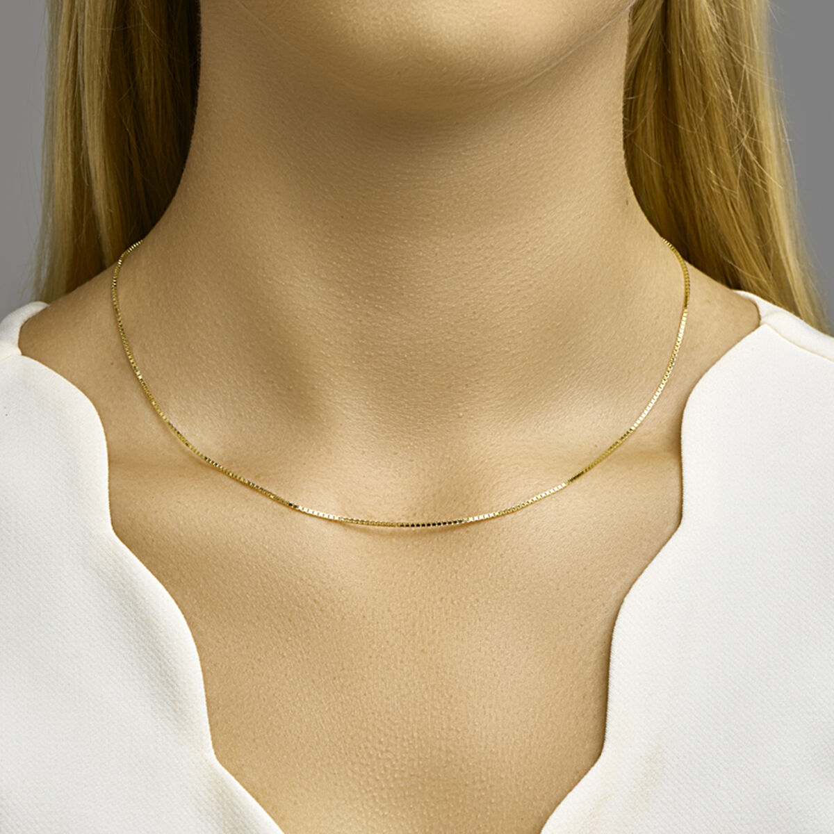 Een vrouw met lang blond haar draagt een Van der Kooij Collectie collier venetiaans 1,1 mm 14K geelgoud en een wit topje met V-hals, waarbij de eenvoudige elegantie van de ketting wordt benadrukt tegen een effen grijze achtergrond.