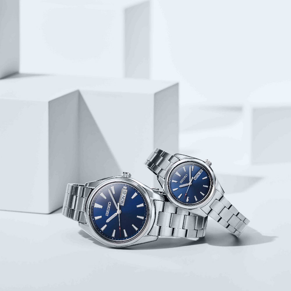 Twee Seiko SUR341P1 horloges met blauwe wijzerplaten en metalen banden staan op witte geometrische blokken. De grotere geeft 10:08 weer; de kleinere 10:12. Deze horloges zijn gemaakt van saffierglas, stralen robuuste betrouwbaarheid uit en hebben een weekdagweergave.