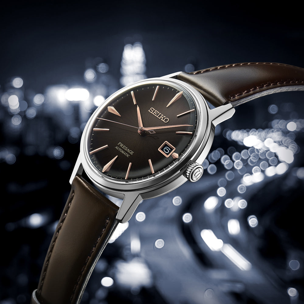 Close-up van de Seiko Presage herenhorloge SRPJ17J1, met een bruine leren band en zwarte wijzerplaat met roségouden indexen en wijzers. Hardlex mineraalglas zorgt voor duurzaamheid, terwijl het wazige stadsgezicht s nachts elegantie toevoegt met witte en gele bokehlichten.