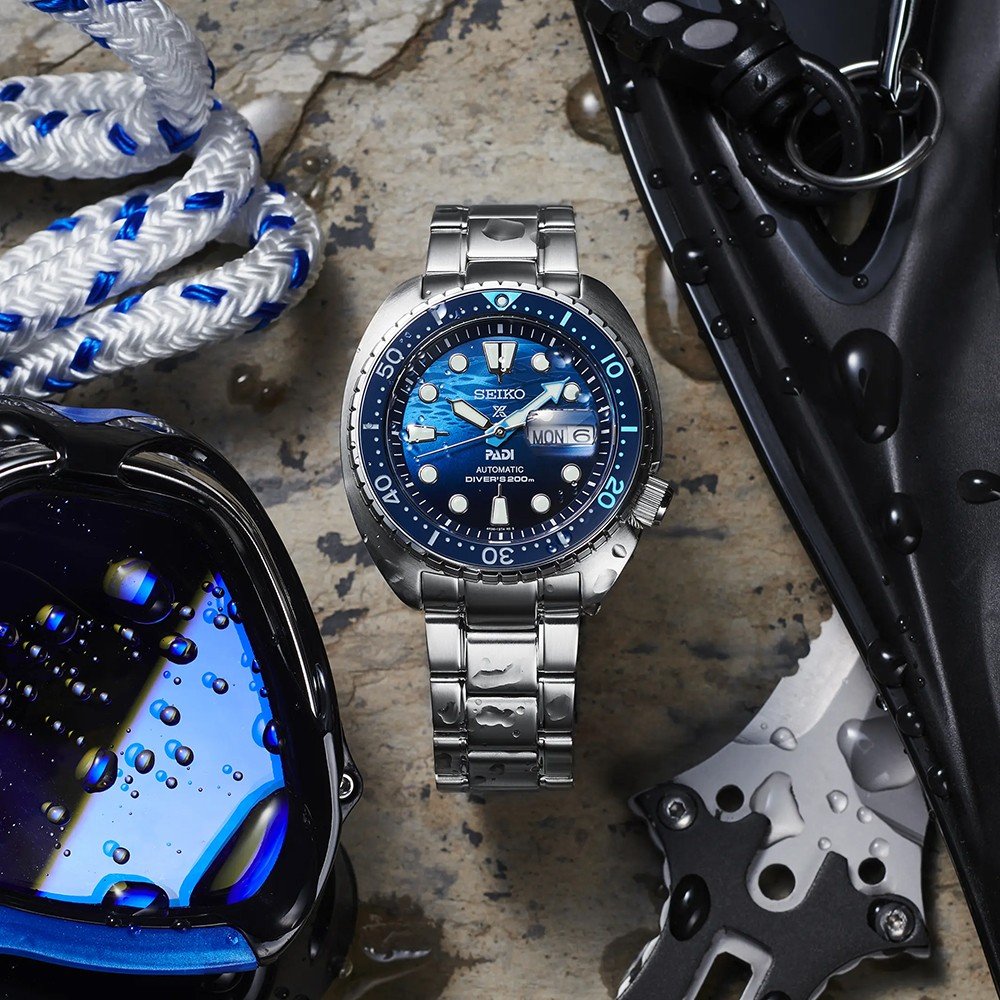 Een zilveren Seiko Prospex herenhorloge SRPK01K1 met een blauw en zwart gezicht en metalen band rust op een rotsachtig oppervlak. In de buurt glinstert duikuitrusting met waterdruppels: een blauw reflecterend masker, gevlochten touw en mes suggereren recente onderwateravonturen.