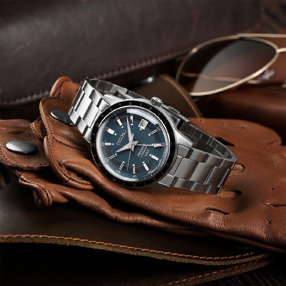 Een Seiko Presage SSK009J1 horloge met een zilveren band en een zwart-blauwe wijzerplaat rust op bruine leren handschoenen, aangevuld met een zonnebril met bruine lenzen. Dit elegante uurwerk, met een automatisch uurwerk, staat tegen een vintage leren achtergrond.